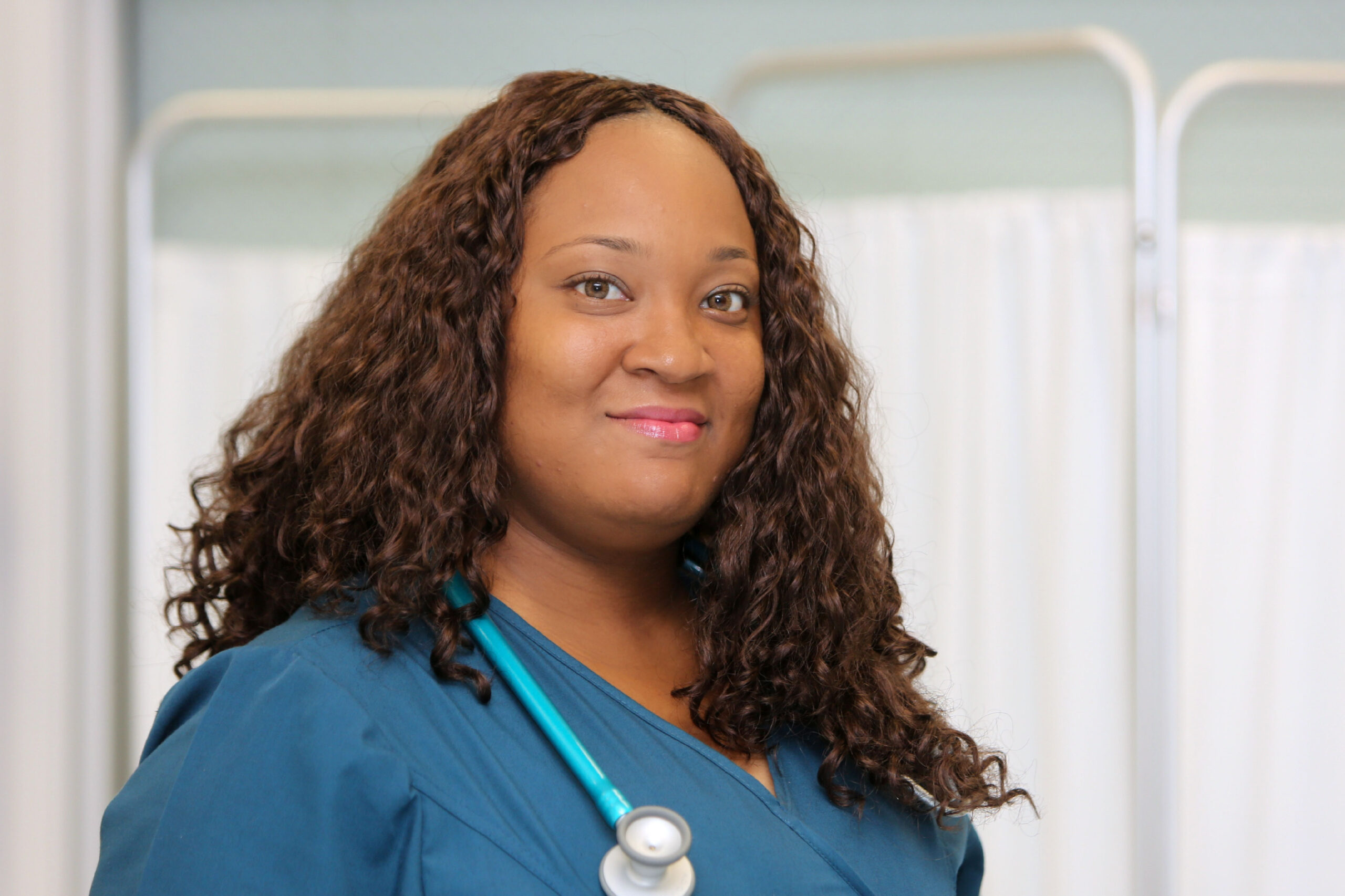 A woman with brown curly hair and brown eyes smiles at the camera, wearing dark blue scrubs and a stethoscope