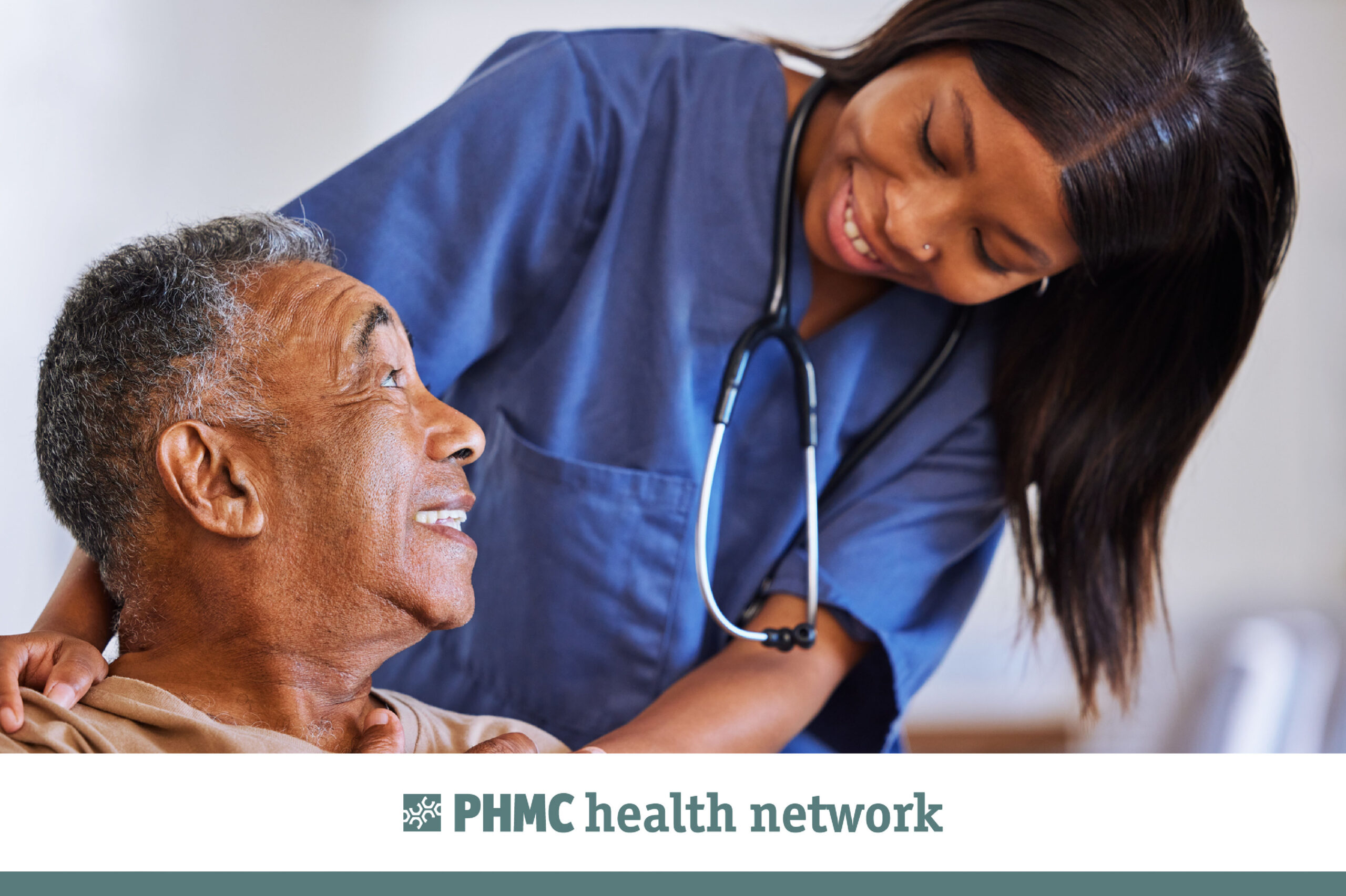 A young woman with brown hair, wearing dark blue scrubs and a stethoscope, smiles and leans over an older man with dark greying hair