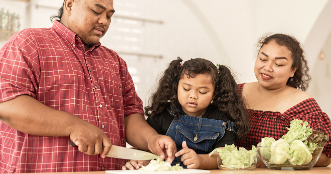 Father cutting vegetables at the kitchen counter as mother and child watch.