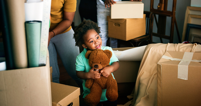 Child holding a teddy bear as his family unpacks boxes in their new house.