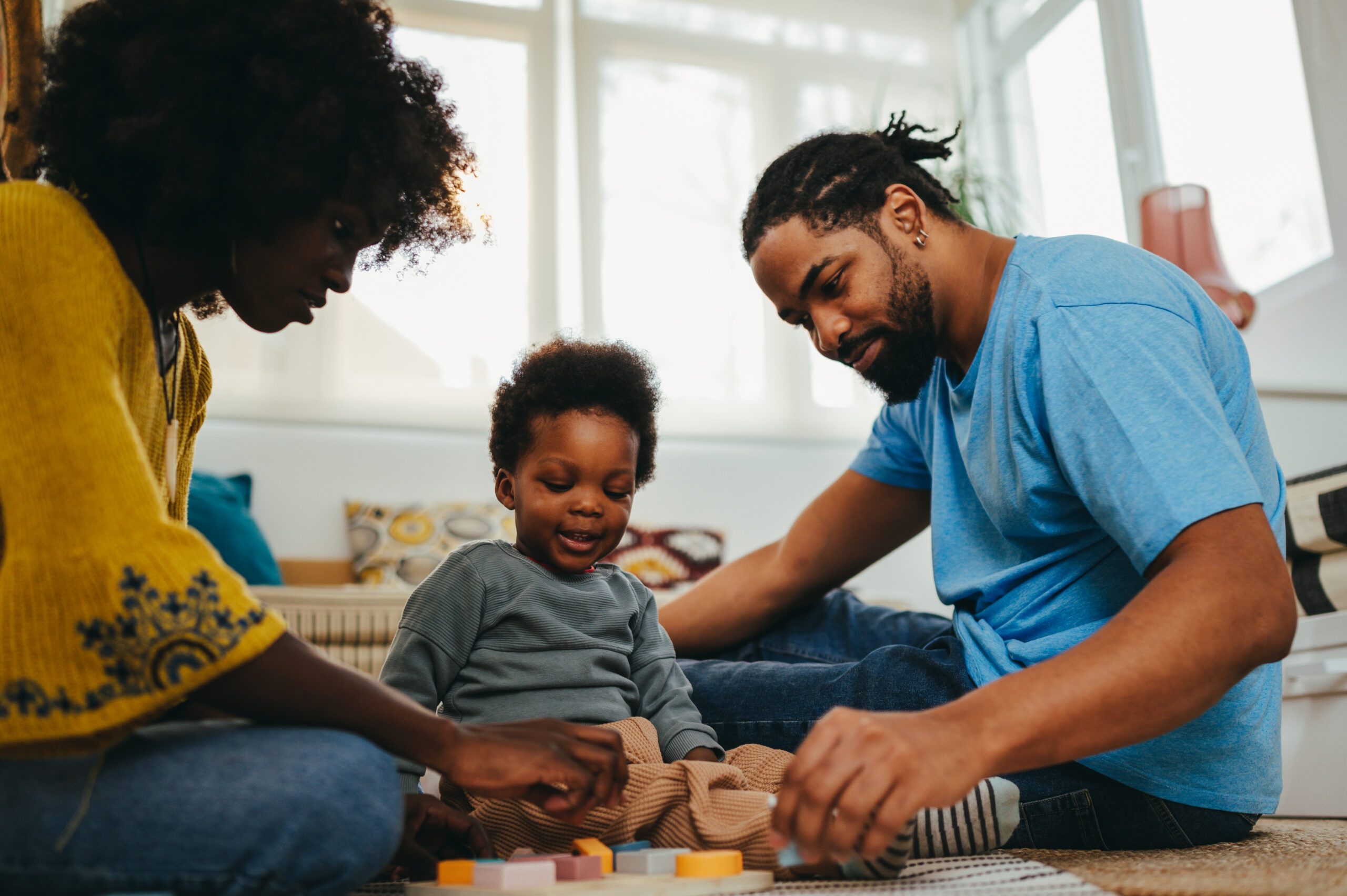 An african american family is having fun with a Montessori toy while playing with a puzzle on the floor in a living room. The diverse parents are teaching their son shapes and colors through the game.